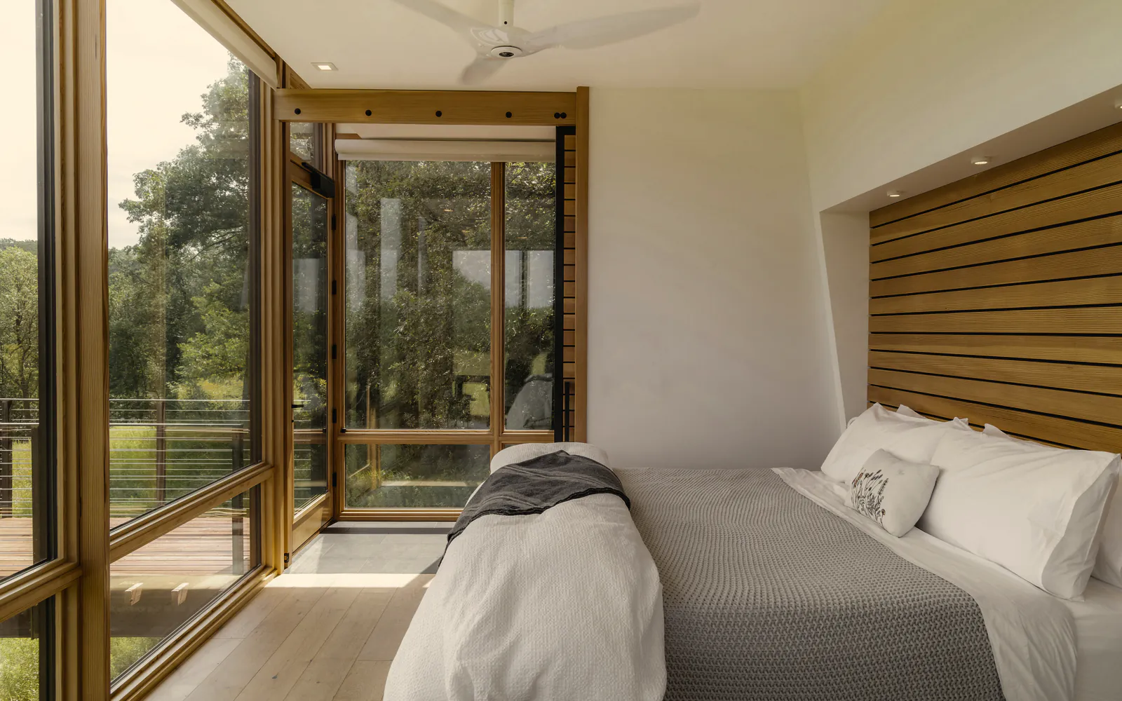 A made bed in white linen under a slatted timber bedhead, with glass on two sides to the trees