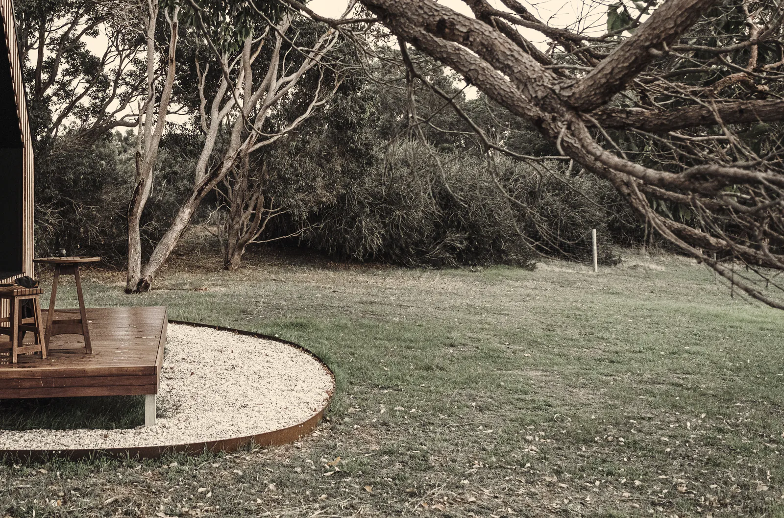 A small timber deck with two stools under a twisted coastal gum, more gums and scrub behind