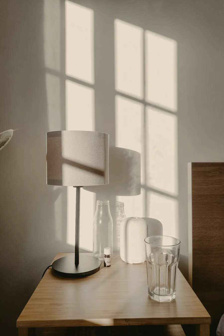 A white lamp and a glass of water on the pale timber bedside, window light on the white wall