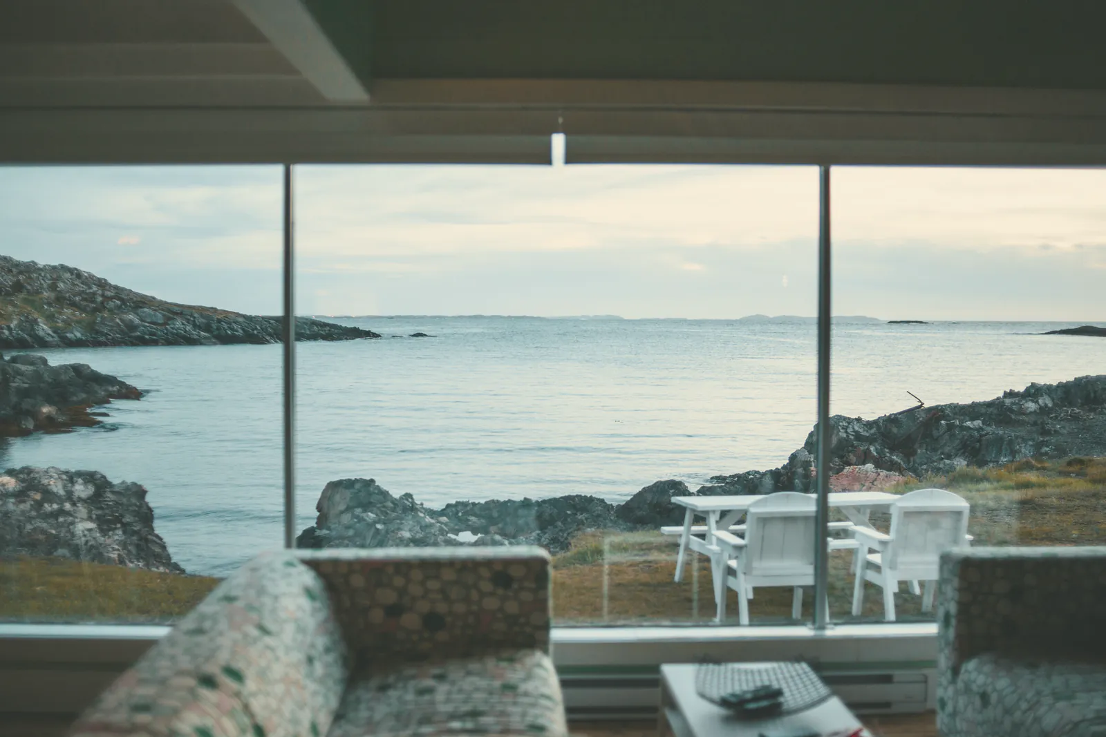 The sitting room’s wall of glass onto a granite cove and a grey sea, two deep sofas in the foreground and a picnic table on the grass outside