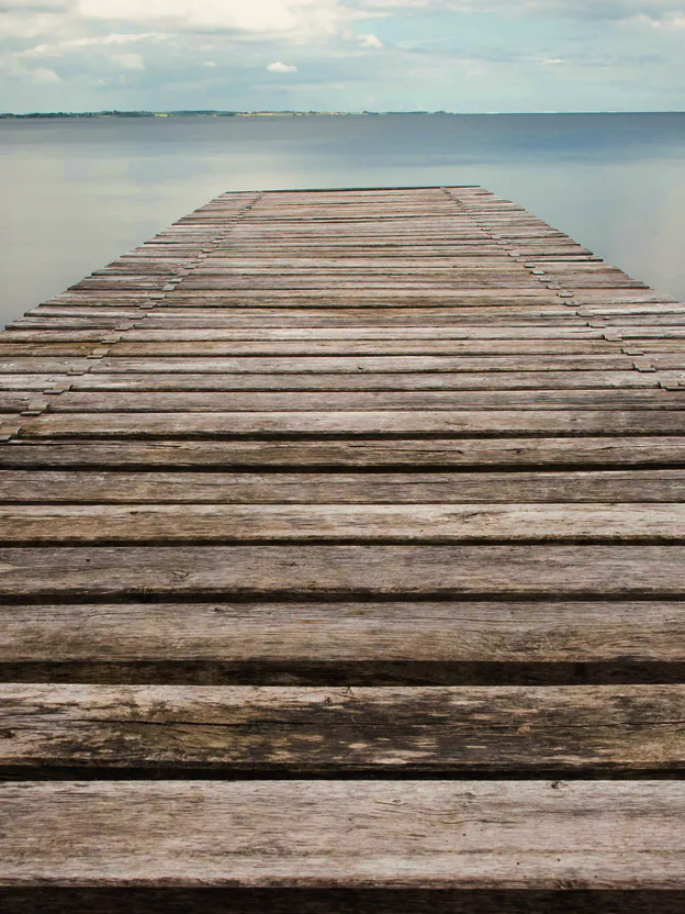 The weathered boards of an old timber jetty running out over silver, calm water