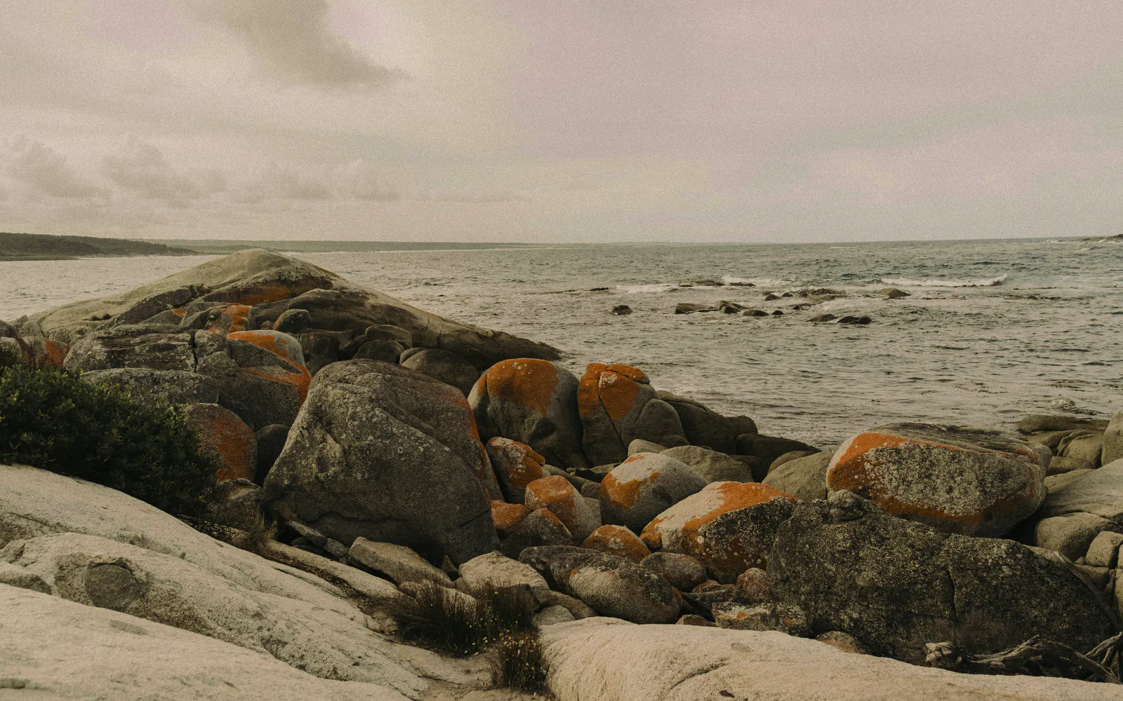 Rounded granite boulders with orange lichen at the edge of the water, the grey bay beyond under a silver sky