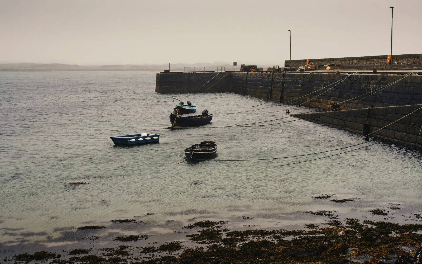 Dinghies on their moorings off the old stone wharf in an overcast calm, kelp along the shore