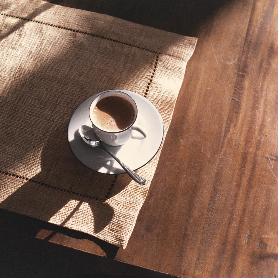 A cup of coffee on a linen runner across the timber table in low morning sun