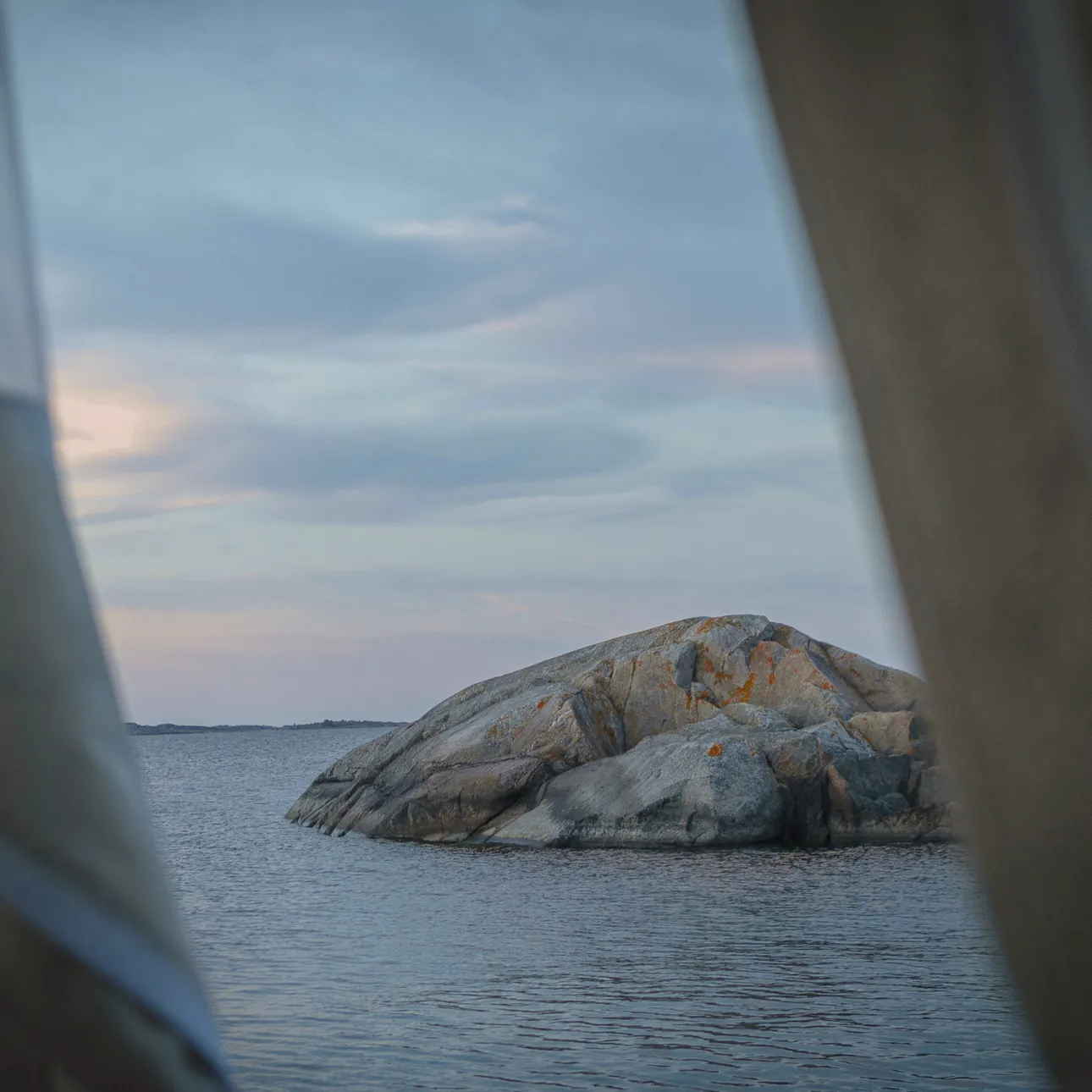 A sheer curtain drawn back from a window onto a calm sea and a granite islet at dusk
