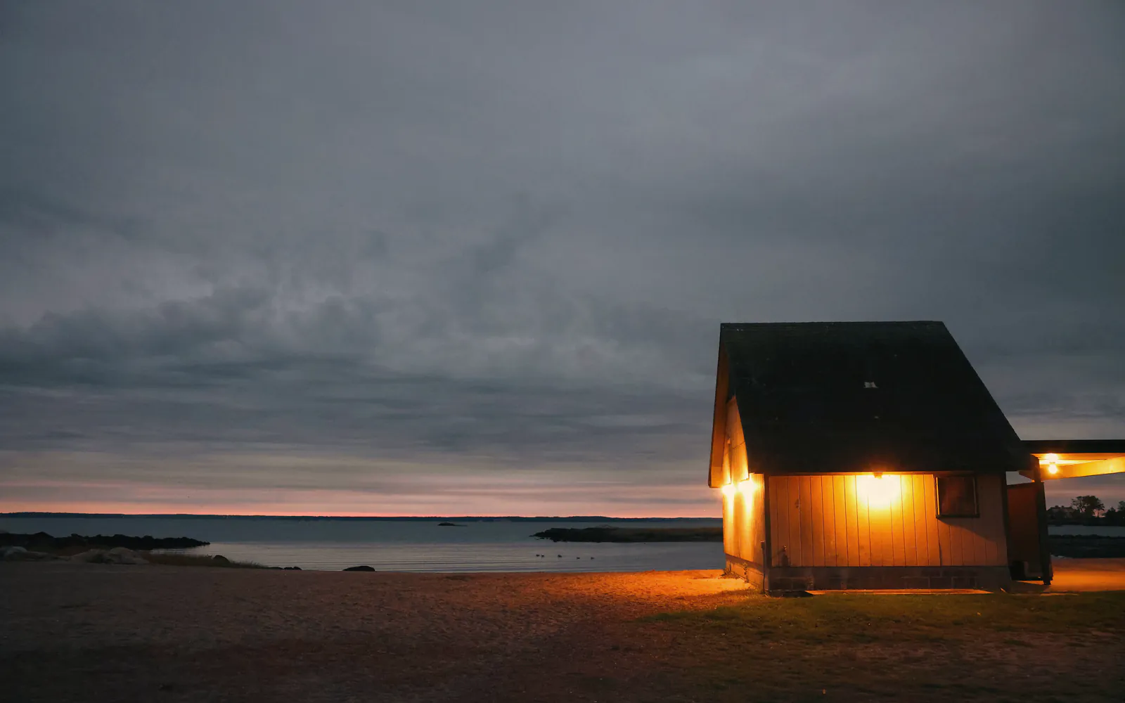 A small timber house lit warm from inside at early dusk, on the sand by a calm sea