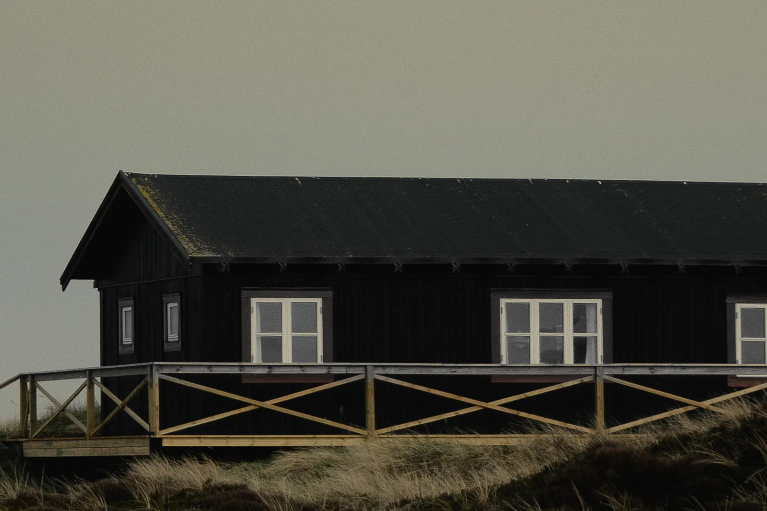The dark board walls and white windows of the house above the timber rail of its deck, dune grass below