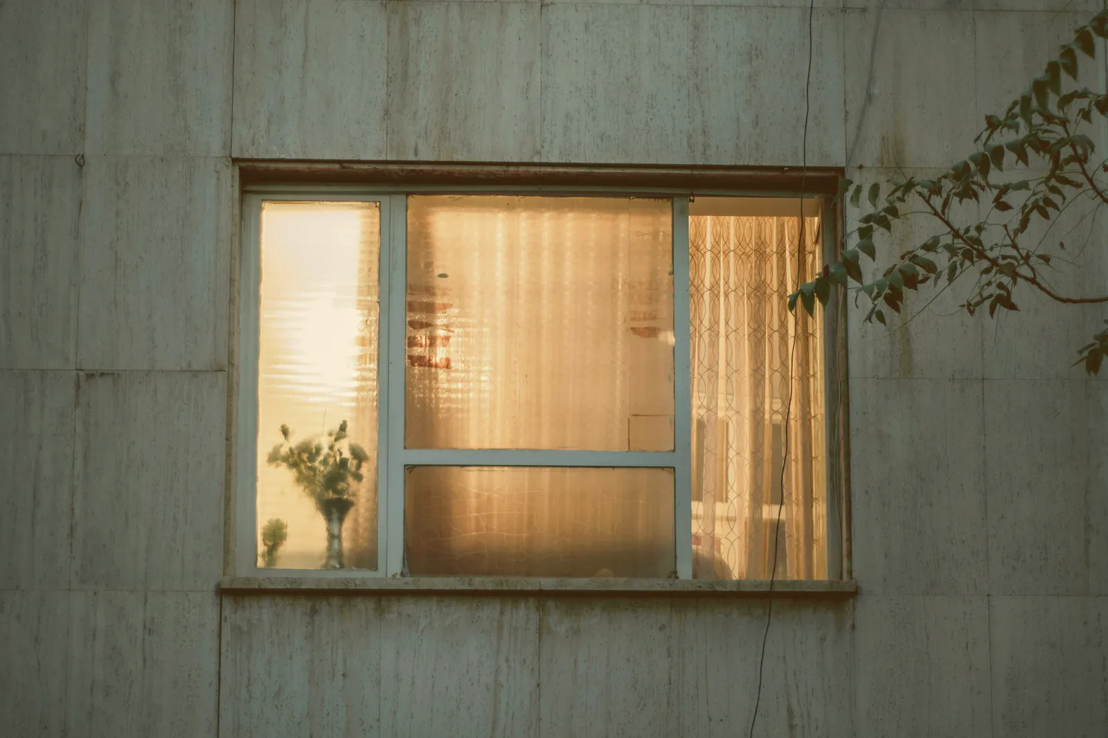 A window lit warm from inside in a weathered board wall at dusk, a sheer curtain and dried flowers on the sill