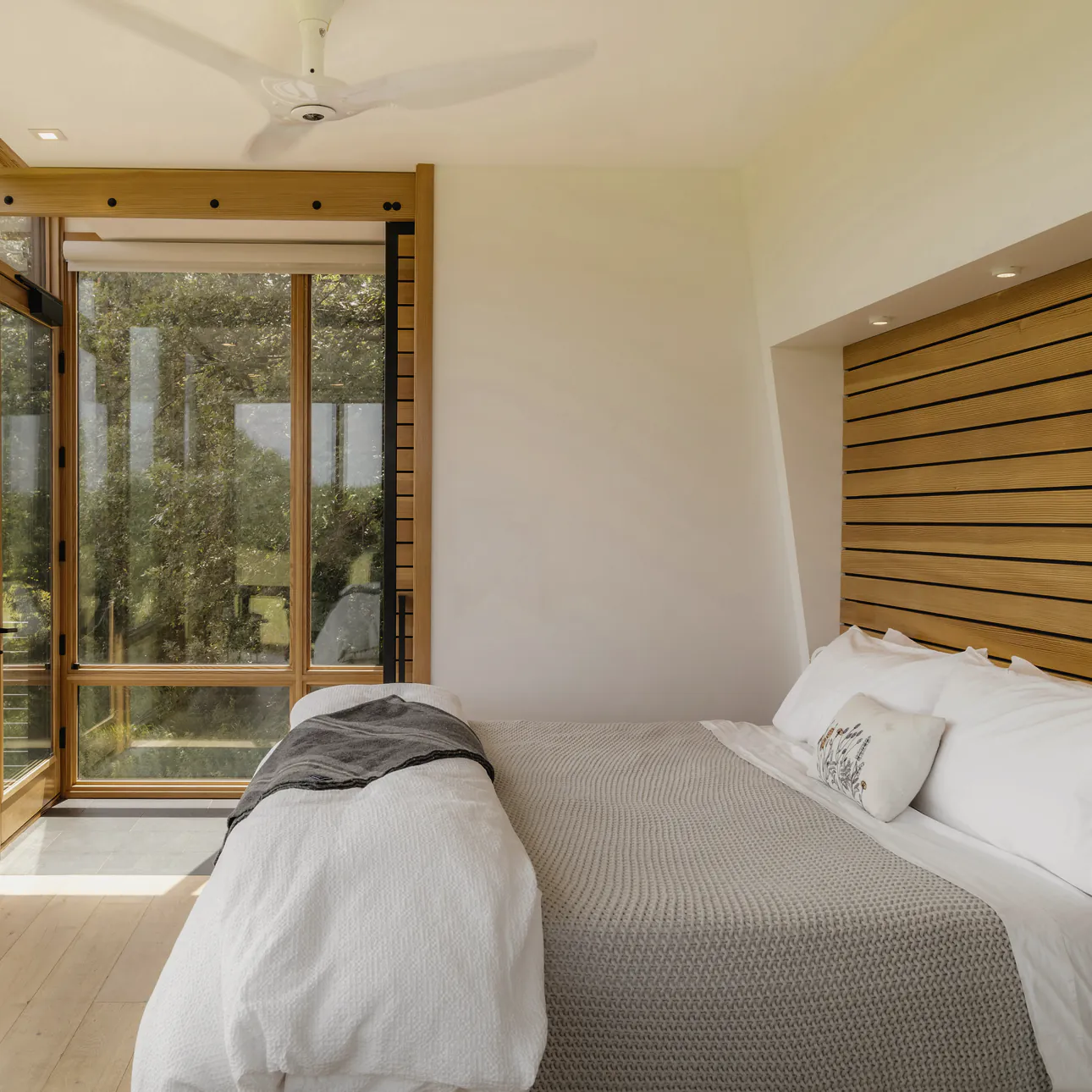 A made bed in white linen under a slatted timber bedhead, with glass on two sides to the trees