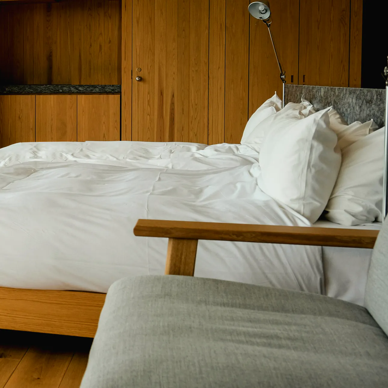 A made bed in white linen on an oak frame against an oak wall, with a reading lamp at the bedhead and a grey armchair at its foot