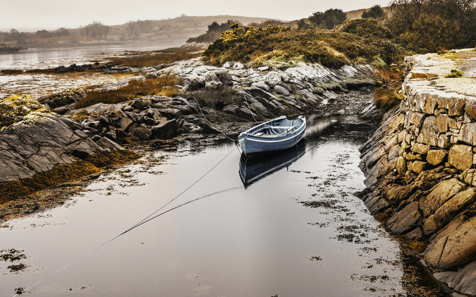 A rowing boat on its lines in a still inlet beside a dry stone harbour wall, gorse and low hills in a sea fog