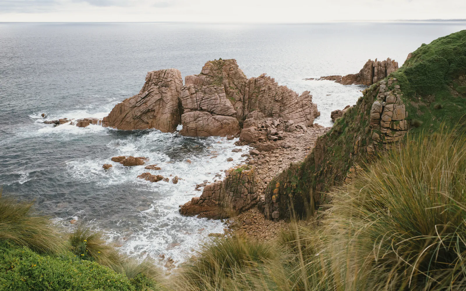Granite stacks and white surf below a slope of tussock on the headland, the sea grey in a soft haze