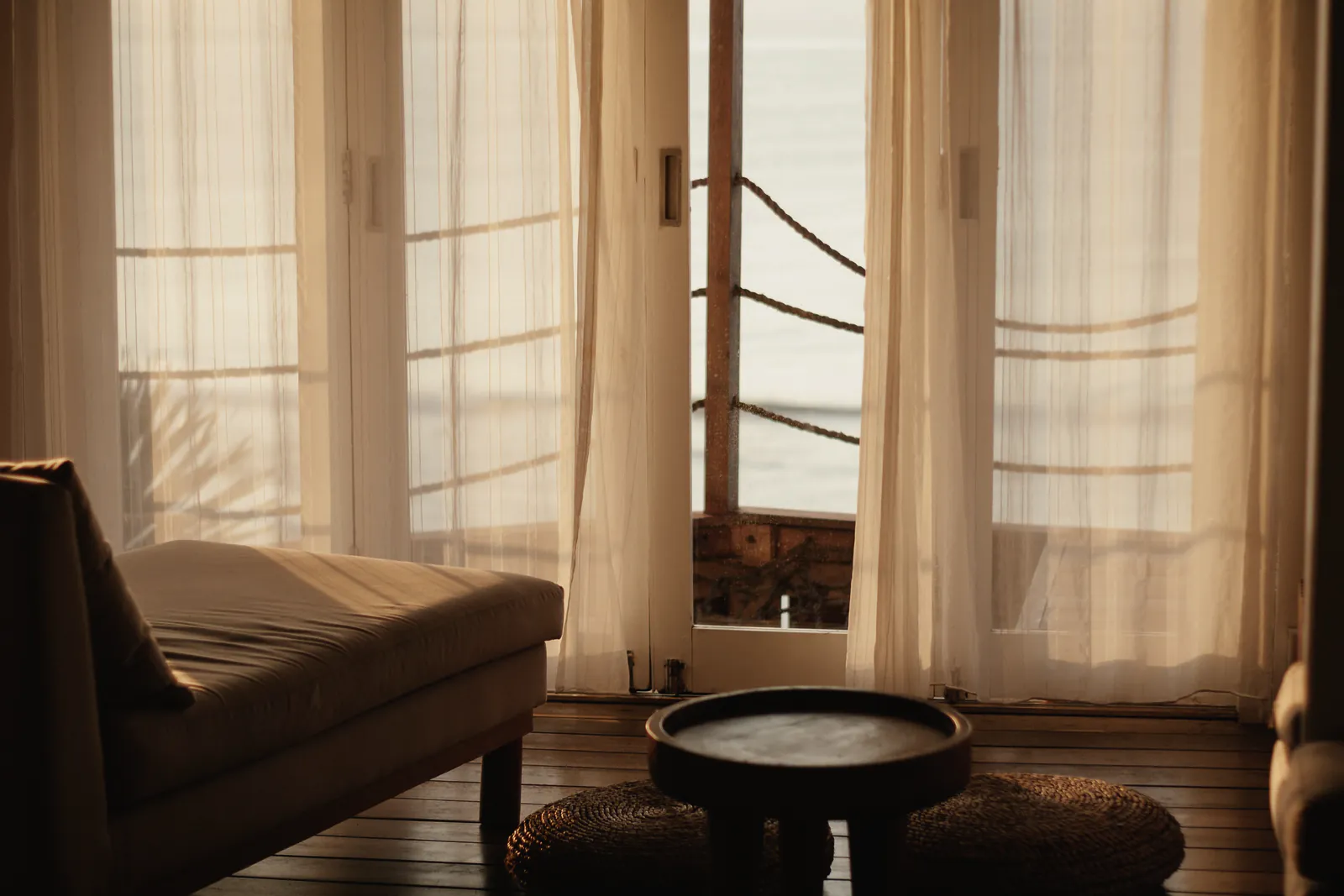 A linen daybed and a round timber table by sheer curtained glass doors open to a deck and the sea, in low gold sun