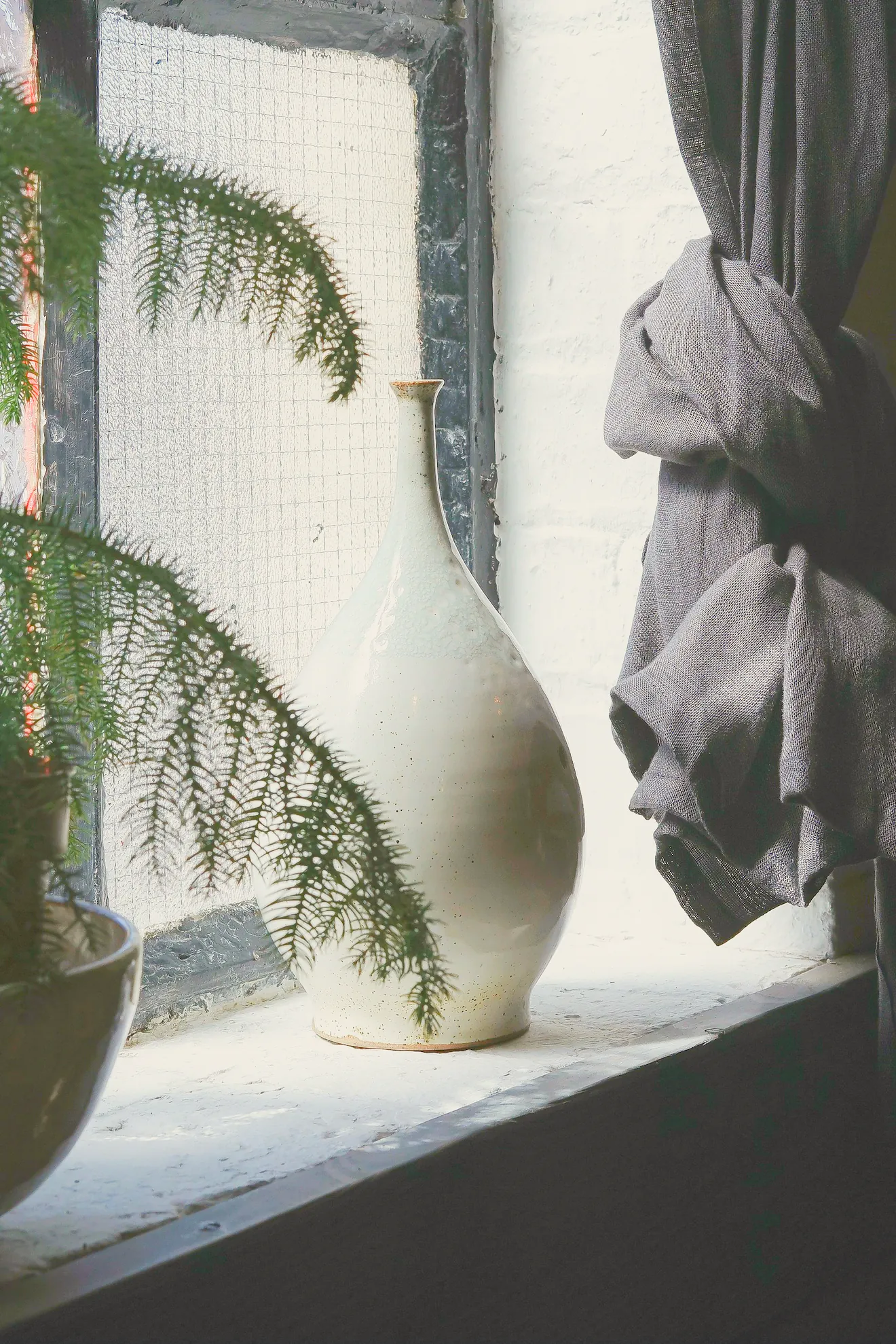 A white stoneware vase on a deep window sill in soft light, a grey linen curtain tied back beside it