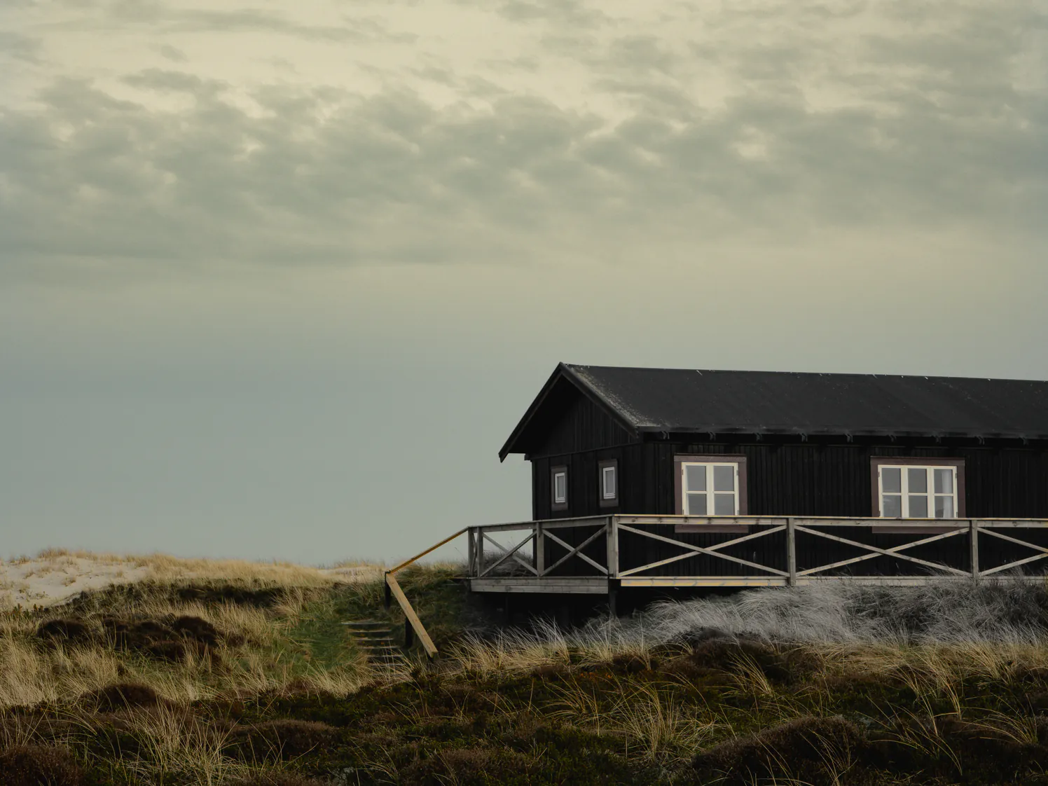 A low dark timber house end on, on its dune under a silver overcast sky, a railed deck along its side and dune grass below