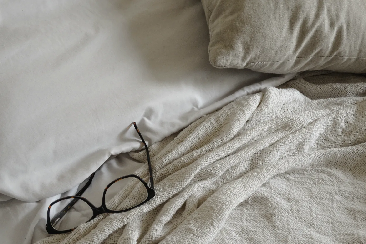 Reading glasses on a cream woven blanket over white linen, a linen pillow at the head of the bed