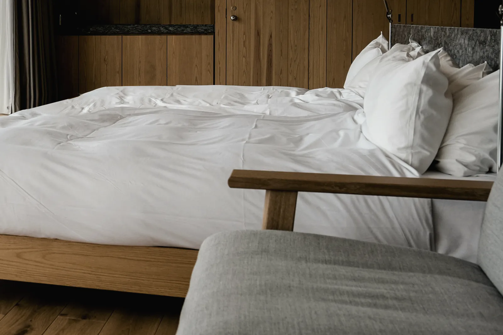 A made bed in white linen on an oak frame against an oak wall, with a reading lamp at the bedhead and a grey armchair at its foot