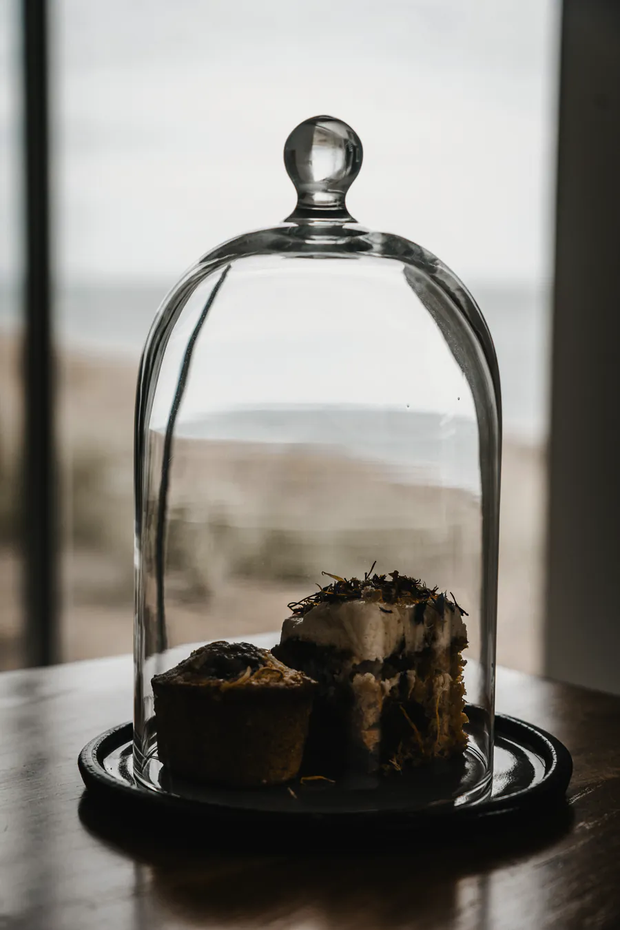 A cake under a glass dome on the timber table, the beach pale through the window behind