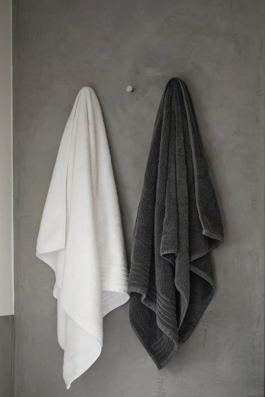 A white towel and a charcoal towel on hooks against the grey stone wall of the bathroom