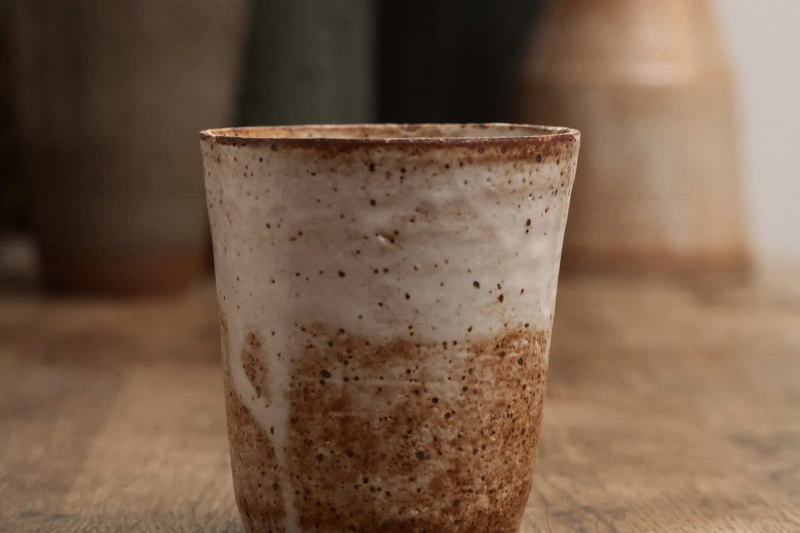 A speckled stoneware cup on the oak table, other pots soft behind it