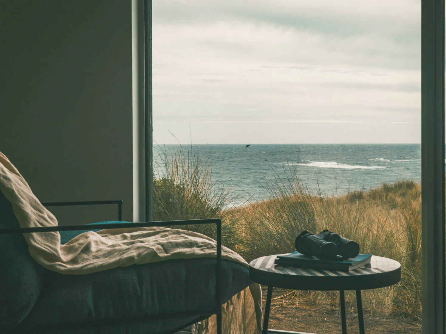 A reading chair with a linen throw at a wide window, binoculars on a side table and dune grass running down to the sea