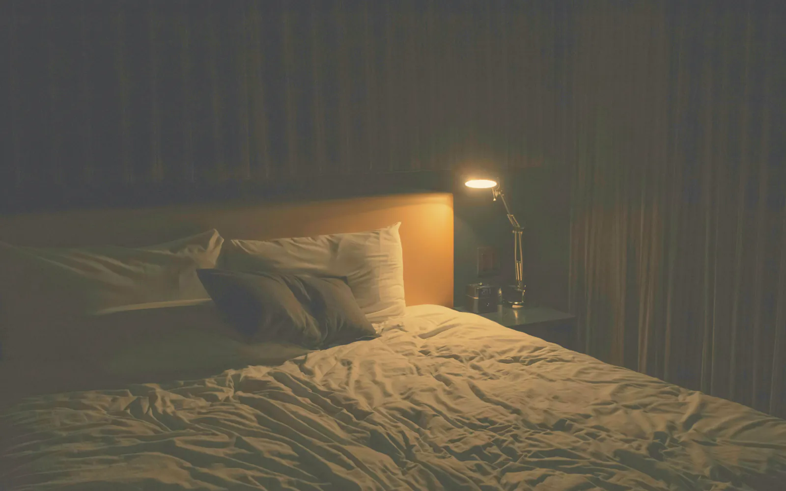 A reading lamp lit over the bed in a dark room at the end of the day