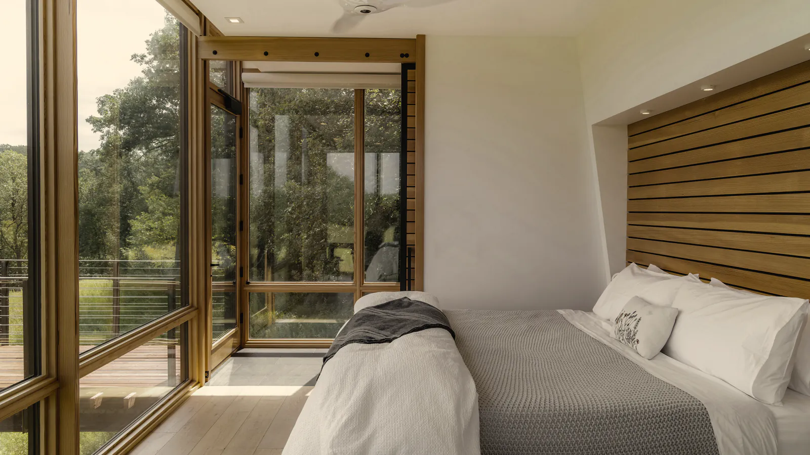 A made bed in white linen under a slatted timber bedhead, with glass on two sides to the trees