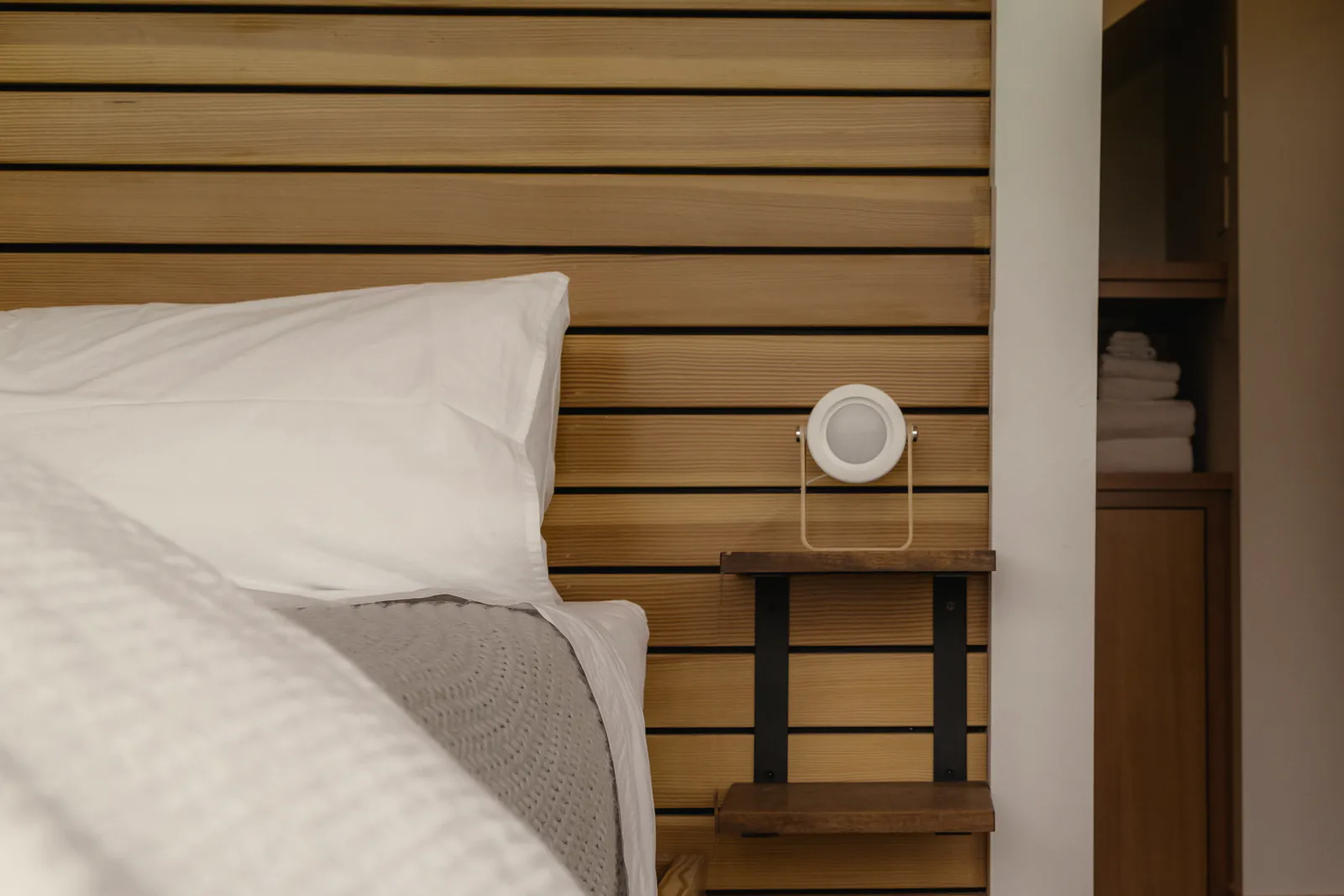 A white pillow under the slatted timber bedhead, a lamp on its shelf and the door to the shower room beyond