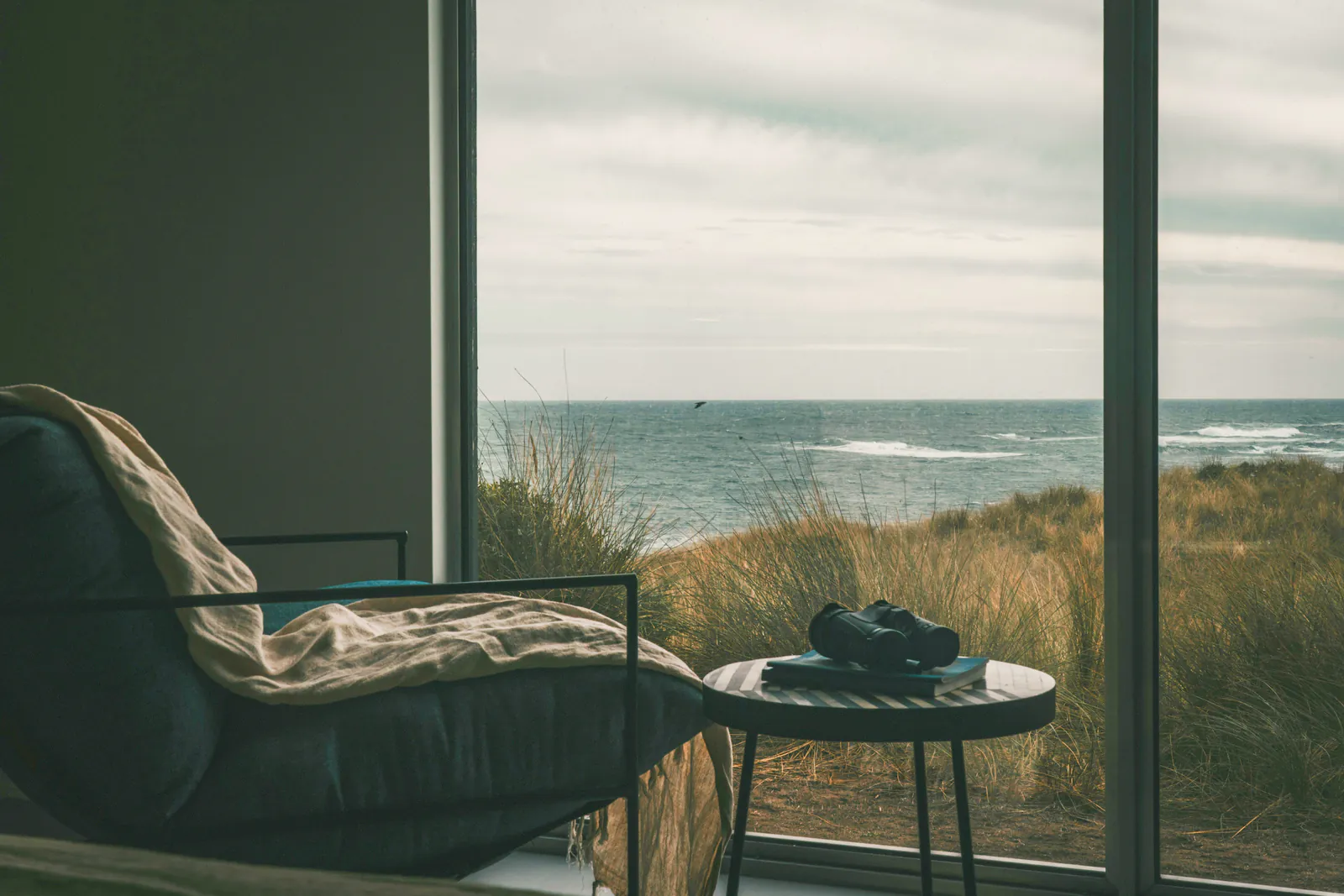 A reading chair with a linen throw at a wide window, binoculars on a side table and dune grass running down to the sea
