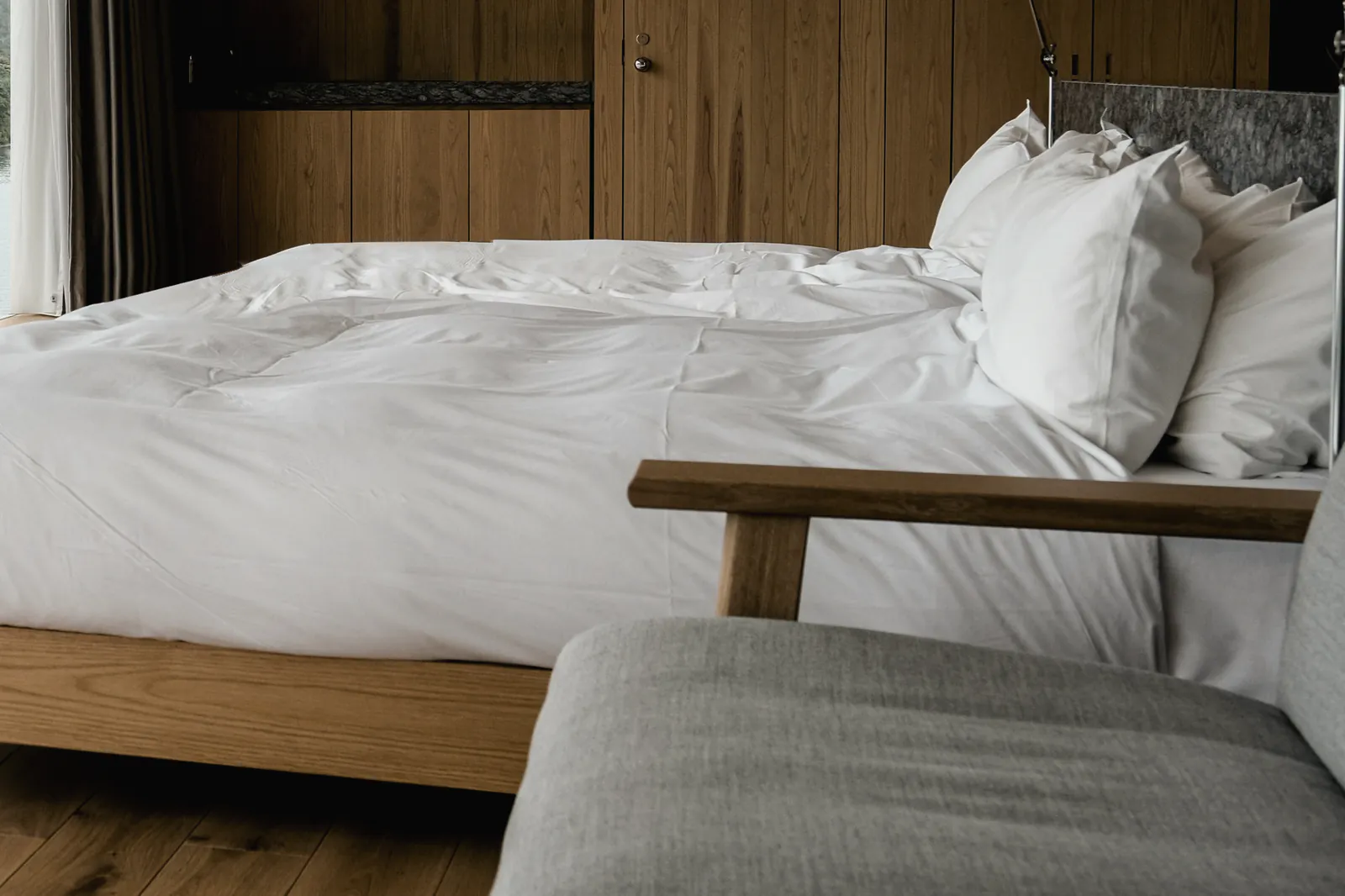 A made bed in white linen on an oak frame against an oak wall, with a reading lamp at the bedhead and a grey armchair at its foot