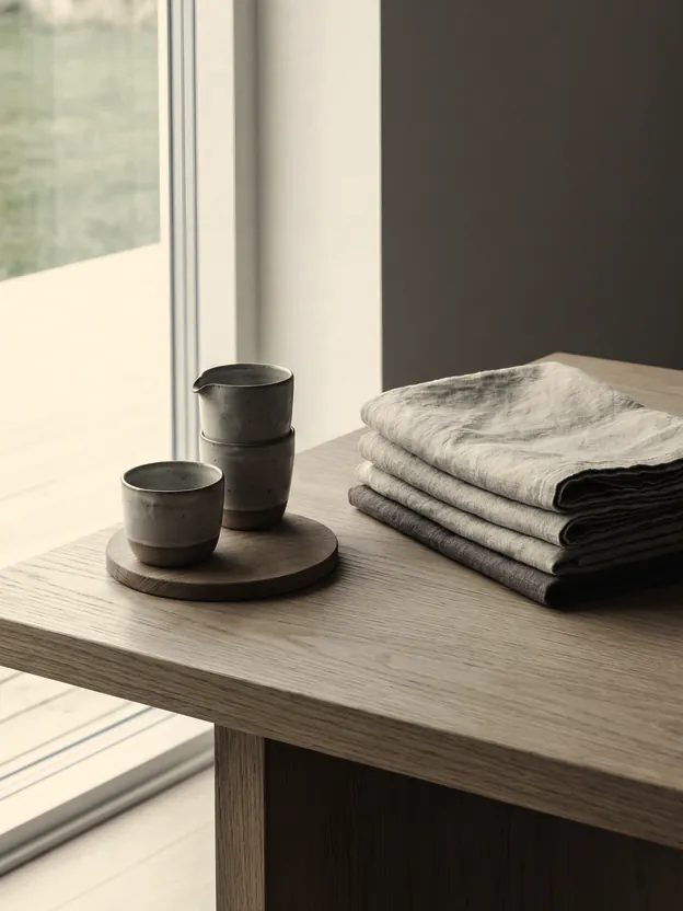 Folded linen on a pale oak table by tall glass, two handmade cups beside it