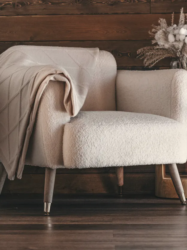 A cream armchair with a knitted blanket against a dark timber wall, dried grasses in a jar beside it
