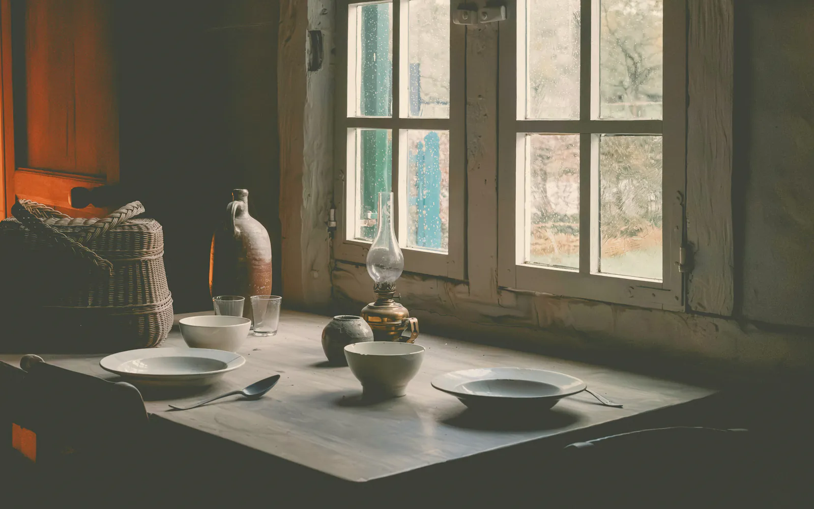 Stoneware bowls laid on the timber table by small paned windows in the early dusk, an oil lamp and a jug beside them