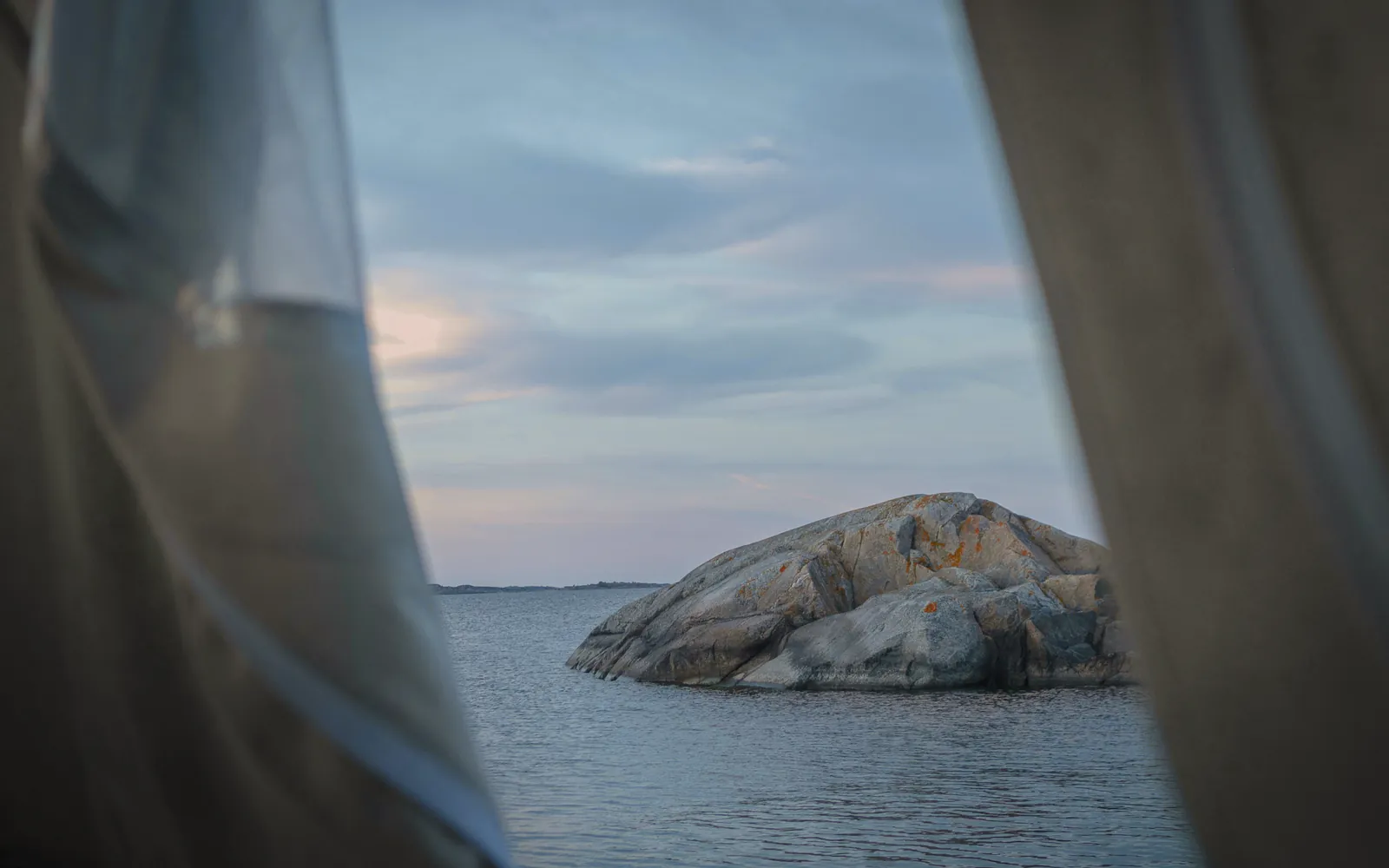 A sheer curtain drawn back from a window onto a calm sea and a granite islet at dusk